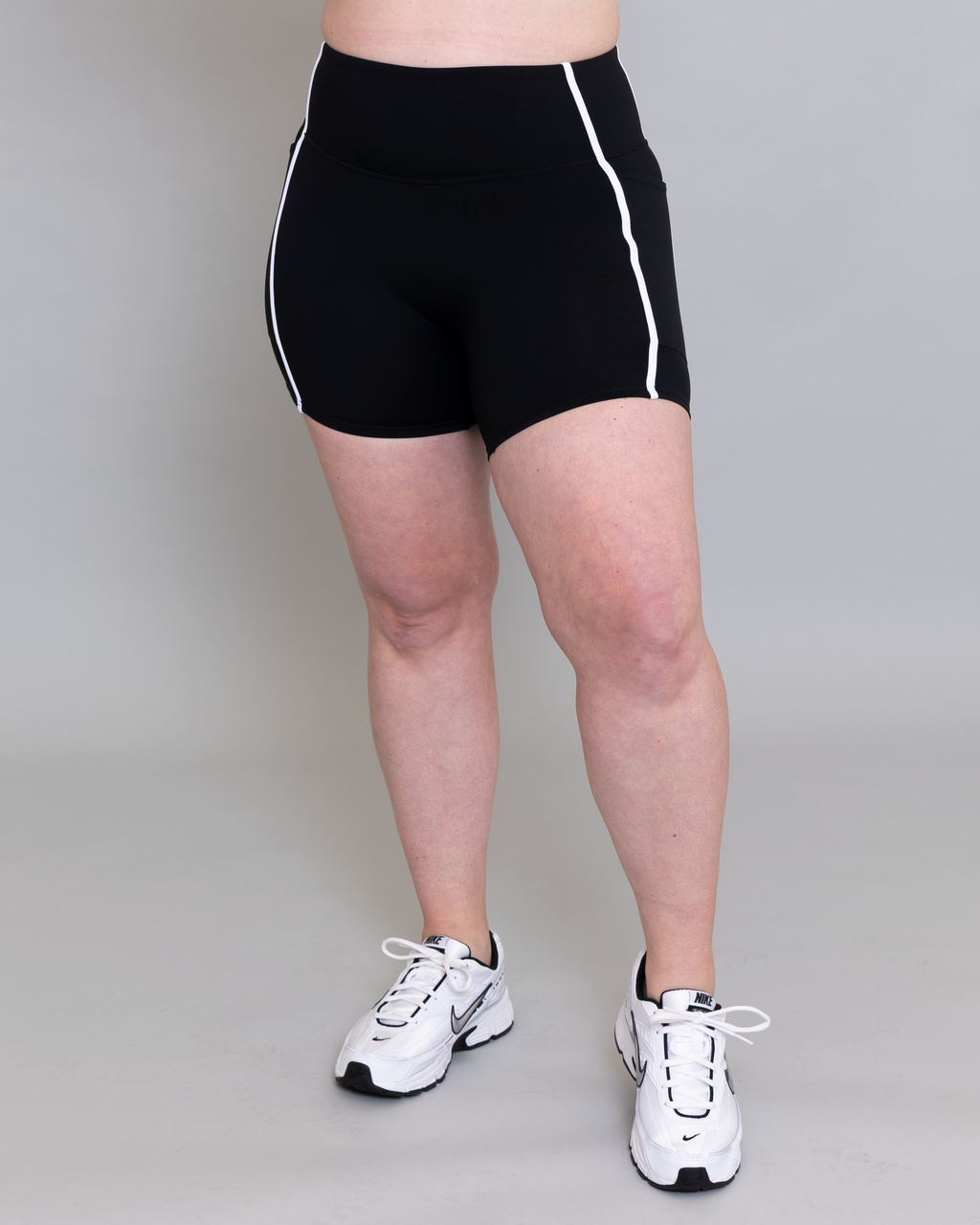 Person wearing black athletic shorts with white stripes and white sneakers on a plain background
