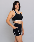 Woman wearing a black sports bra and shorts with white trim on a gray background
